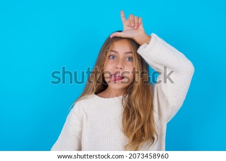 brunette kid girl wearing knitted sweater over blue background gestures with finger on forehead makes loser gesture makes fun of people shows tongue