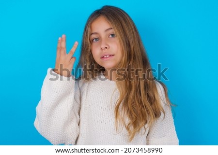 brunette kid girl wearing knitted sweater over blue background smiling and looking friendly, showing number three or third with hand forward, counting down