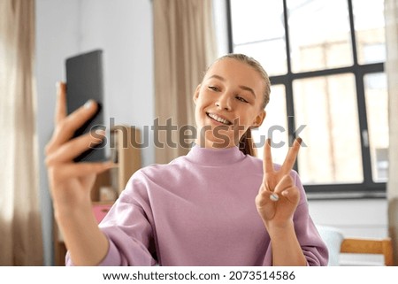 people, children and technology concept - happy smiling teenage girl with smartphone taking selfie and showing peace gesture at home
