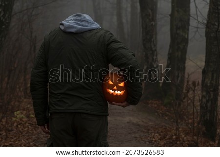 Headless man holds Jack O'Lantern Halloween pumpkin head with orange glowing carved face under his arm and stands on countryside road in autumn foggy forest. Copy space. Halloween costume theme.