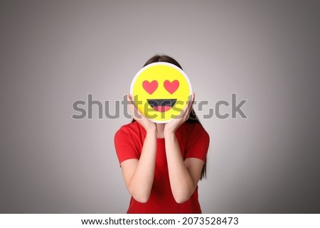 Woman covering face with heart eyes emoji on grey background Royalty-Free Stock Photo #2073528473