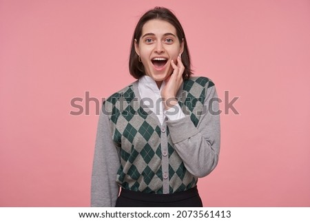 Indoor shot of young brunette female wears stylish touching her cheek, keeps mouth opened widely with surprised facial expression.