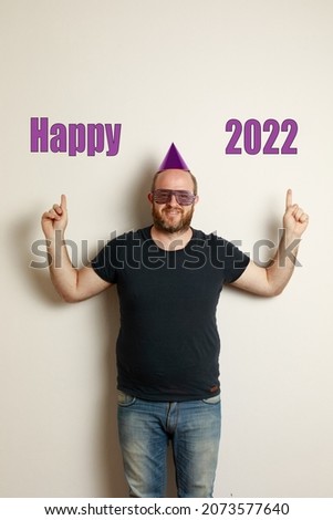 A Funny image of Caucasian man with a birthday hat and funny glasses; points to a text Happy 2022. Neutral background.