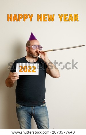 A Humorous image of Caucasian man celebrating the new year with a birthday hat, funny glasses and a bugle. It has a sign with 2022. Neutral background with Happy new year writing.