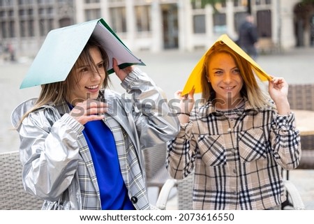 Two female students sitting outside at a terrace table covering their heads for the rain with two colored plastic folders. High quality photo