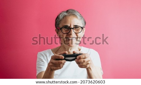 Old senior woman smiling to camera while holding a console pad, video games playing pastel pink removable background, video games old people, copy space, white shirt space