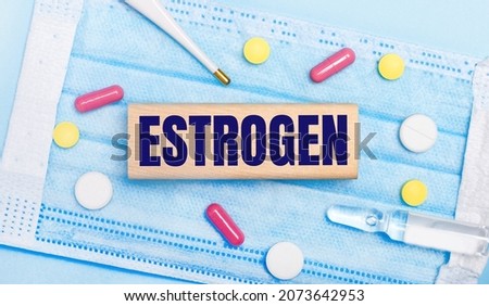 On a light blue disposable face mask there are tablets, a thermometer, an ampoule and a wooden block with the text ESTROGEN. Medical concept