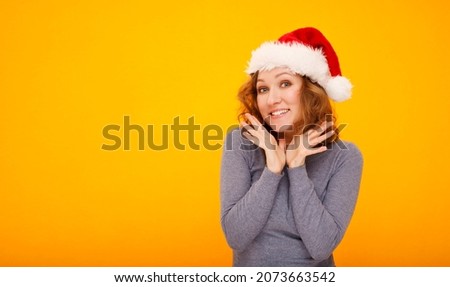 Young happy woman with curly hair in christmas hat standing over yellow background smiling with happy face looking at camera with hands near face. Place for your text.