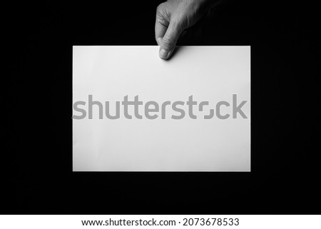 B+W image of male hand holding blank sheet of paper against black background with space for copy.