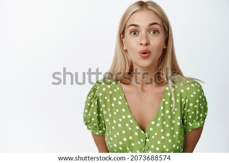 Portrait of cute blond girl looks curious, pucker lips and looking intrigued, standing in green dress over white background
