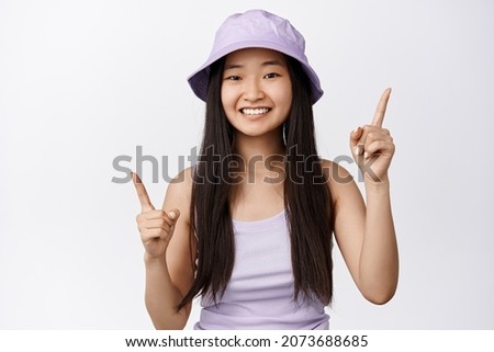 Smiling asian girl pointing fingers sideways, showing left and right, two options, standing in tank top and bucket hat against white background