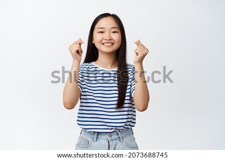 Cute korean girl shows finger hearts gesture and smiling, optimistic and positive face expression, happy attitude, standing over white background