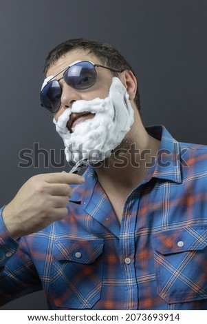 A man in sunglasses, a plaid shirt and a foam beard shaves with a razor