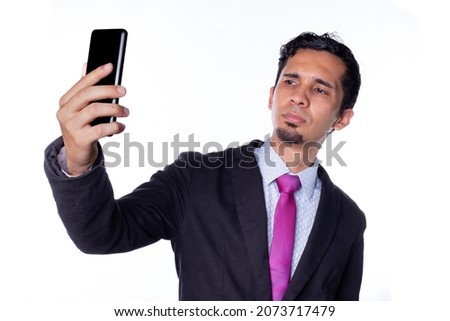 Latin businessman taking a selfie. Young adult brown skinned man using smart phone to take a selfie, isolated on all white background.