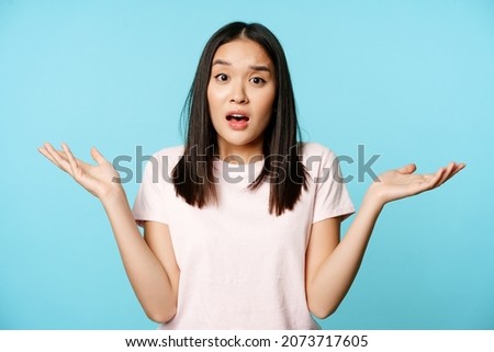 Confused asian girl spread empty hands sideways, shrugging shoulders clueless, standing in tshirt over blue background Royalty-Free Stock Photo #2073717605