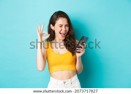 Technology and lifestyle concept. Cheerful brunette female model say yes, winking and showing okay sign, holding mobile phone, standing over blue background