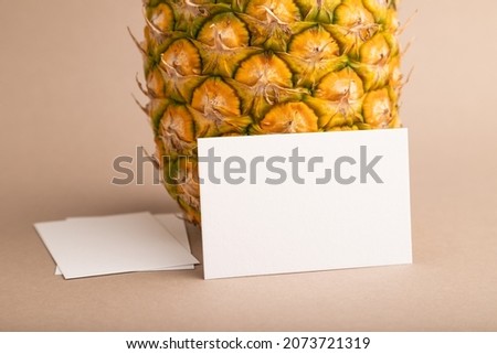 White paper business card with ripe pineapple on orange pastel background. Side view, copy space. Tropical, healthy food, vacation, holidays concept.