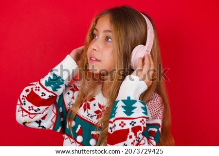 brunette kid girl in knitted sweater christmas over red background  wears stereo headphones listens music concentrated aside. People hobby lifestyle concept