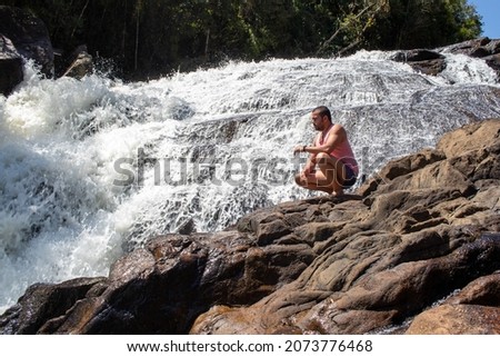 waterfall, man relaxing on a sunny day, sitting on the rock at the edge of the waterfall, in the background beautiful landscape and blue sky