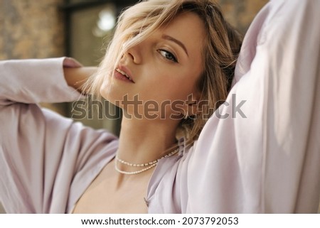 Close-up of young caucasian woman looking seductively into camera, covering half of face. Blue-eyed beauty with short hair holds hands behind head, wears earrings in ears and chain on neck. Royalty-Free Stock Photo #2073792053