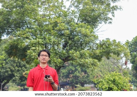 Portrait of thoughtful Asian man in red polo shirt standing in the park while enjoying music using an earphone and holding mobile phone