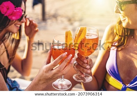 Two girlfriends toasting sitting on chaise lounge on the beach at sunset - best friends talking together and drinking on summer - closeup picture, focus on the glasses