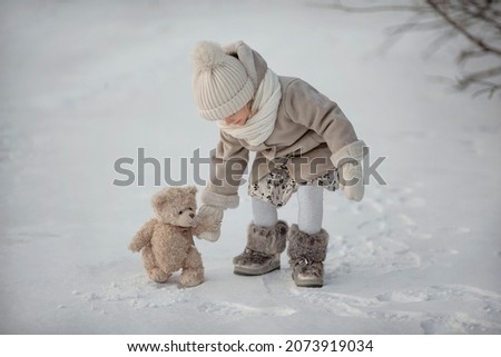 Happy child girl playing with teddy on a winter walk in nature. 
Outdoor fun for winter vacation. Baby playing with teddy on the snow-covered lake. Image with selective focus