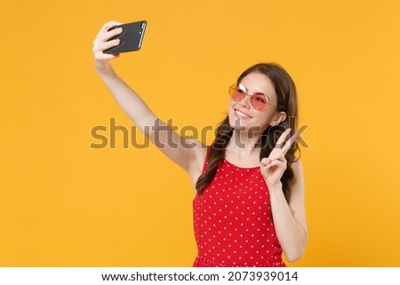 Smiling brunette woman girl in red summer dress, eyeglasses posing isolated on yellow background. People lifestyle concept. Mock up copy space. Doing selfie shot on mobile phone showing victory sign