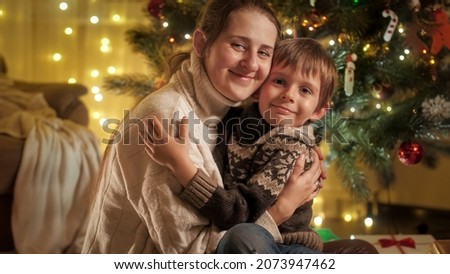 Portrait of cheerful smiling boy with mother hugging next to Christmas tree and looking in camera. Families and children celebrating winter holidays