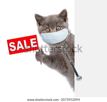 Kitten wearing stethoscope and medical protective face mask looks from behind empty banner and shows sales symbol. Isolated on white background 
