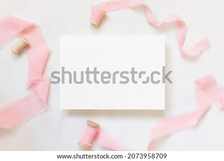 Paper card on white marble table with dried pink flowers and silk ribbons top view. Flat lay with horizontal blank card. Romantic invitation or greeting card mockup, copy space