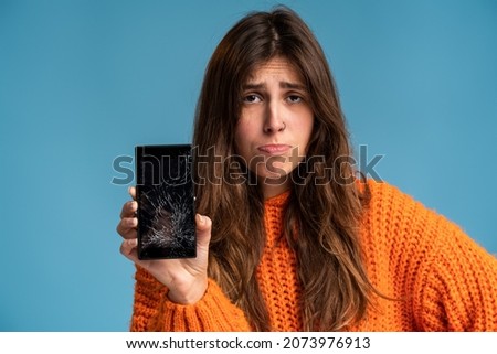 Portrait of a pretty sad woman in orange jumper holding smartphone with broken screen standing over blue background and looking at the camera 