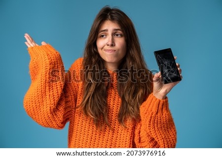 Portrait of a pretty sad woman in orange jumper holding smartphone with broken screen and throwing up hands in disbelief over blue background 