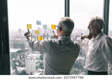 Picture of young business man talking to his older business partner. They are in white shirt and black tie. They are sitting on a table in a hotel lobby. 