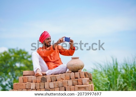 Indian farmer sitting on bricks and using smartphone at agriculture field.