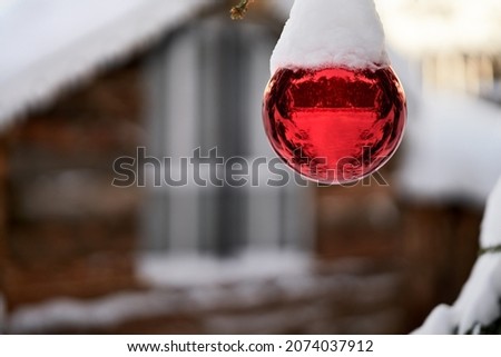 A red glass ball under the snow on a branch of a Christmas tree on the background of a blurry image of a chalet. Selective focus. Copy space.                               