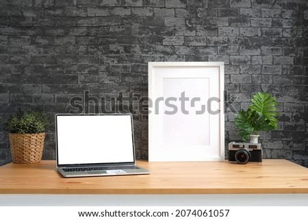 Laptop computer, empty picture frame, camera and houseplant on wooden table with brick wall.