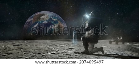 Astronaut sits on his knee on the lunar surface and pours blue earth's soil through his fingers. Planet Earth rise at the horizon. Elements of this image furnished by NASA. Royalty-Free Stock Photo #2074095497