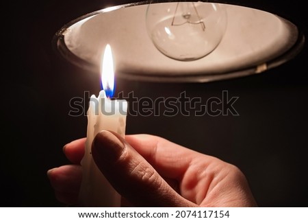 A woman's hand with burning candle in complete darkness looking on a switched off light bulb at home. Blackout, electricity off, energy crisis or power outage, concept image. Royalty-Free Stock Photo #2074117154