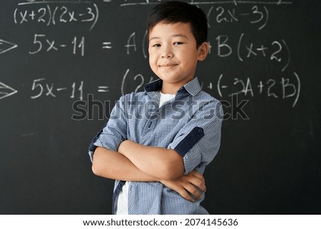 Happy cheerful proud confident smiling cute asian schoolboy with arms crossed standing posing in classroom on chalkboard blackboard with formulas background. Elementary preteen school kid portrait. Royalty-Free Stock Photo #2074145636