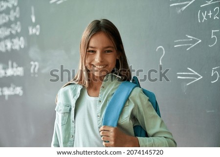Happy cheerful smiling cute hispanic schoolgirl with backpack standing posing in classroom on chalkboard blackboard background. Elementary preteen school kid portrait. Back to school concept.