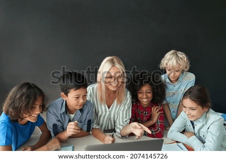 Happy cheerful female teacher in glasses sit together with elementary middle diverse schoolchildren at desk on chalkboard background using looking at laptop computer. Education technologies concept.