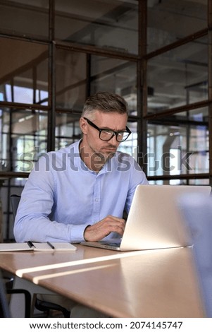 Serious concentrated European Caucasian broker trader in glasses looking at pc laptop working at desk in office. Focused male ceo manager leader analysing financial data using computer. Vertical. Royalty-Free Stock Photo #2074145747