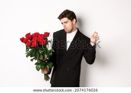 Image of elegant and sassy man in black suit, looking confident and holding bouquet of red roses, going on a romantic date, standing against white background
