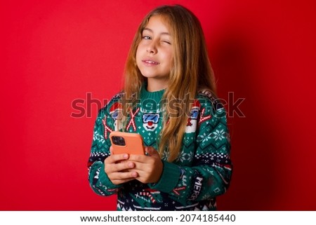 Pleased brunette kid girl in knitted sweater christmas over red background using self phone and looking and winking at the camera. Flirt and coquettish concept.