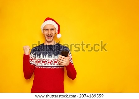 excited man in New Year sweater, red Christmas hat, isolated yellow background with free space for text of advertisement, with phone in his hands shows victory sign. concept - winning, sale, discounts