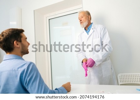 View from back of sitting male patient to mature adult urologist putting pink protective gloves in hands before examining client. Doctor wearing protective glove for rectal examination of man. Royalty-Free Stock Photo #2074194176