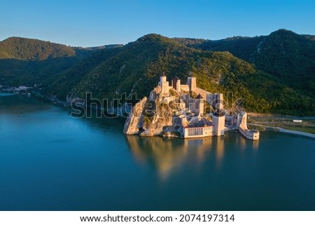 The medieval fortress of Golubac, mirroring in the waters of the Danube. Fortress towers illuminated by warm sunset light, blue sky. Aerial shot. Famous tourist place, Serbia. Royalty-Free Stock Photo #2074197314