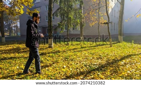 Man in the yard in the fall on a walk controls a quadcopter drone on the street plays and shoots a video side view