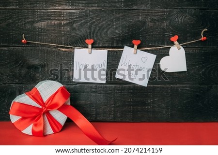 Sticky notes with hearts hang on a wooden wall. Below is a gift box with a bow on a red background. Copy space.